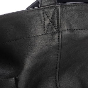 May include: A black leather tote bag featuring a smooth texture, visible stitched seams, and a reinforced handle attachment point with a distinct X-shaped stitch pattern. The bag has a structured top edge and a vertical seam running down the front.