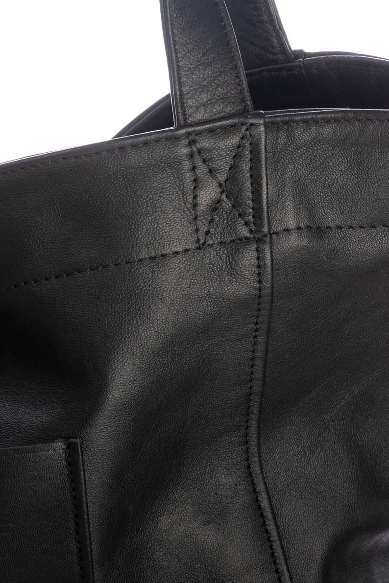 May include: A black leather tote bag featuring a smooth texture, visible stitched seams, and a reinforced handle attachment point with a distinct X-shaped stitch pattern. The bag has a structured top edge and a vertical seam running down the front.