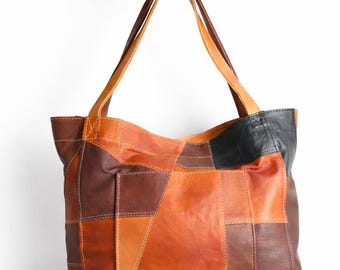 May include: A patchwork leather tote bag in shades of brown, orange, and black. The bag features a rectangular shape with a front pocket and two long handles. The bag is held up by a hand.