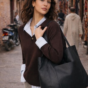 May include: A woman wearing a brown sweater over a collared shirt, carrying a large black tote bag with a front pocket. The background shows a narrow street with buildings and a parked motorcycle.