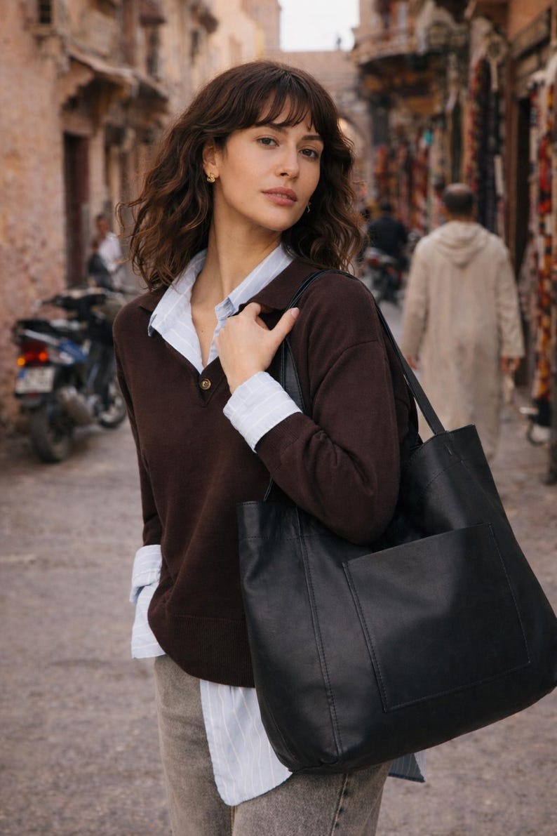 May include: A woman wearing a brown sweater over a collared shirt, carrying a large black tote bag with a front pocket. The background shows a narrow street with buildings and a parked motorcycle.
