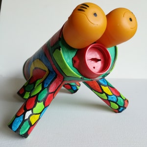 Puede incluir: Una alcancía artesanal caprichosa con forma de criatura con grandes ojos naranjas. El cuerpo es un cilindro pintado con patrones coloridos y abstractos. Las patas también están pintadas con un patrón de escamas en rojo, amarillo y azul.