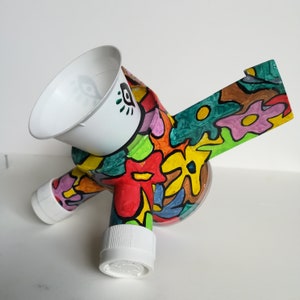 Puede incluir: Una escultura caprichosa hecha a mano, que presenta una taza blanca con un diseño de ojo, conectada a un cuerpo con un patrón floral colorido. La escultura está hecha de materiales reciclados, con una base de plástico transparente y pies de tapón blanco.