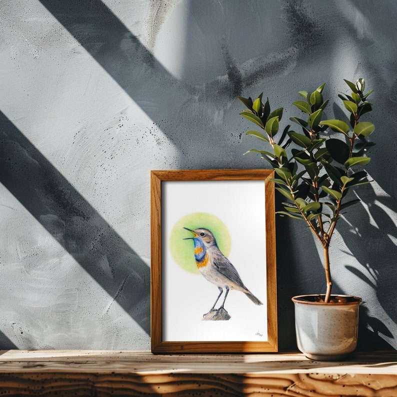 May include: A framed watercolor print of a bird with a blue throat and chest, perched on a rock. The bird is set against a green circular background. The wooden frame sits on a wooden surface, with a small potted plant to the right.