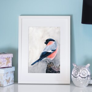 Op de afbeelding: Een ingelijste aquarel van een vogel met een zwarte kop, grijze rug en rode borst. Het kunstwerk wordt tentoongesteld aan een witte muur naast een zwarte lamp en decoratieve dozen. De vogel zit op een tak.