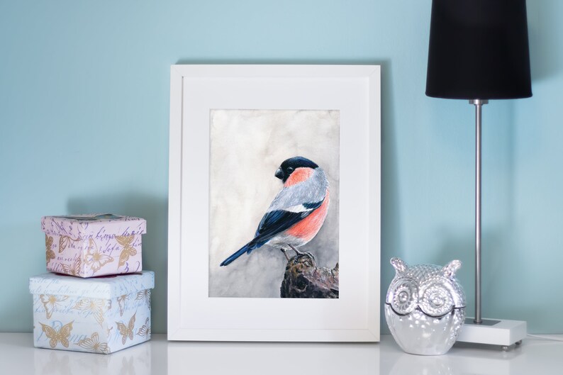 Op de afbeelding: Een ingelijste aquarel van een vogel met een zwarte kop, grijze rug en rode borst. Het kunstwerk wordt tentoongesteld aan een witte muur naast een zwarte lamp en decoratieve dozen. De vogel zit op een tak.