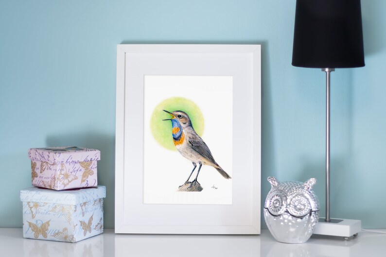 May include: A framed watercolor print of a Bluethroat bird with a green background. The bird has a blue throat, orange chest, and gray and brown feathers. The artwork is displayed on a white table with decorative boxes and a lamp.