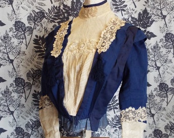 May include: A vintage blue and cream blouse with lace detailing and tassels. The blouse is on a white mannequin and a wooden stand.