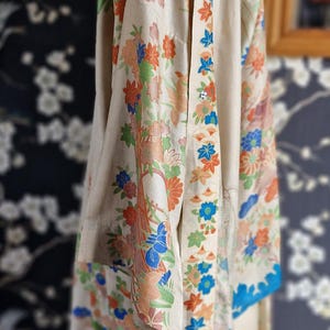 May include: A cream-colored kimono with a floral pattern in orange, blue, and green. The kimono has a blue border and is displayed on a mannequin. The background features a dark floral wallpaper.