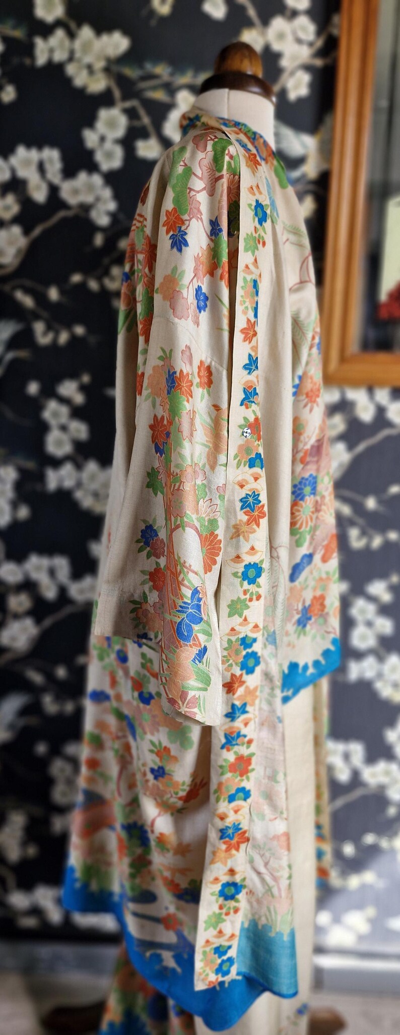 May include: A cream-colored kimono with a floral pattern in orange, blue, and green. The kimono has a blue border and is displayed on a mannequin. The background features a dark floral wallpaper.