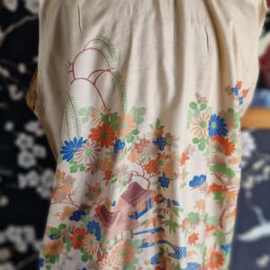 May include: Cream-colored kimono with a colorful floral and landscape design. The pattern includes orange, blue, and green flowers, trees, and buildings. The neckline and hem are trimmed with blue fabric.