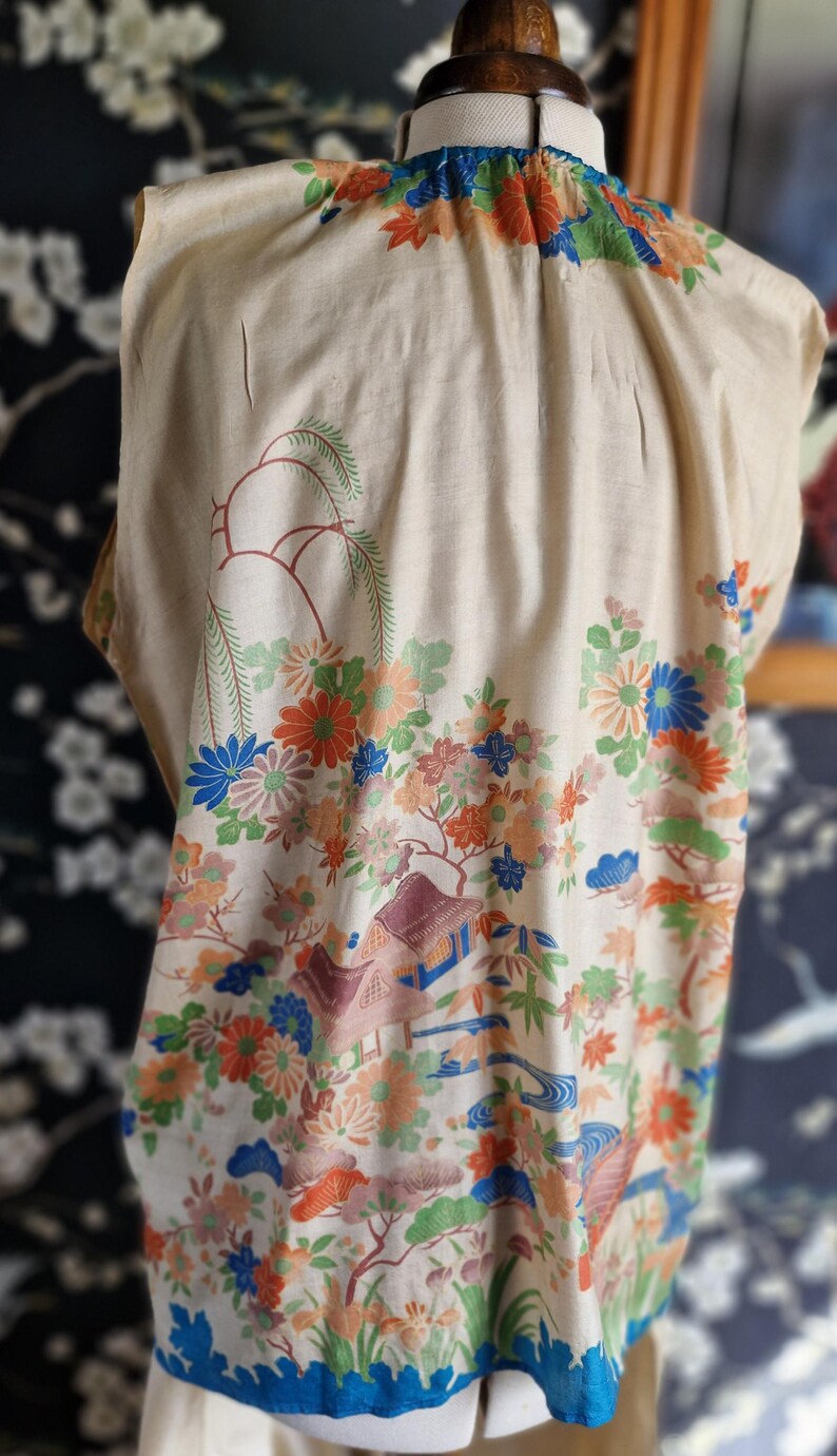 May include: Cream-colored kimono with a colorful floral and landscape design. The pattern includes orange, blue, and green flowers, trees, and buildings. The neckline and hem are trimmed with blue fabric.