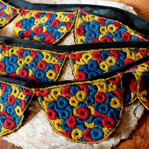 May include: A close-up of a colourful embroidered trim with a scalloped edge. The trim features a repeating pattern of small circles in shades of red, blue, and yellow. The trim is sewn onto a black ribbon.