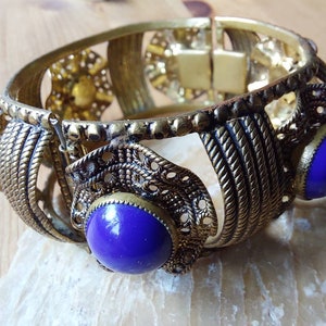 May include: A gold-toned bangle bracelet with intricate detailing and three large, round, purple stones.