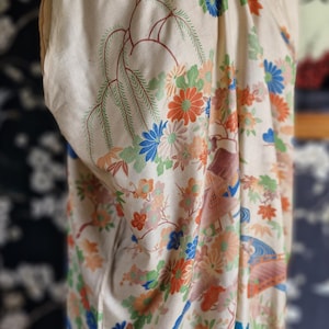 May include: Cream-colored vintage kimono with a vibrant floral pattern in orange, blue, and green. The design includes stylized flowers and branches, with a blue trim along the edges. The kimono is displayed on a mannequin.