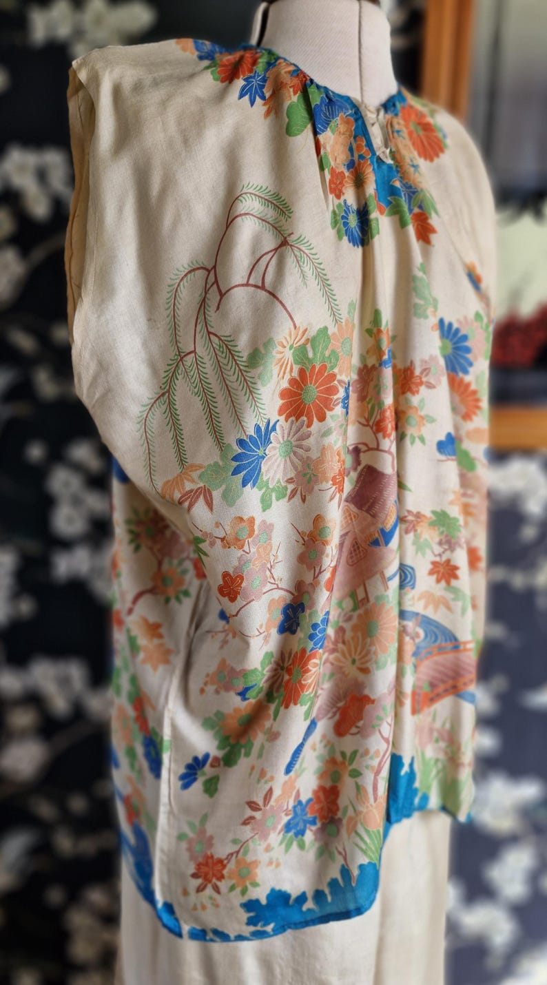 May include: Cream-colored vintage kimono with a vibrant floral pattern in orange, blue, and green. The design includes stylized flowers and branches, with a blue trim along the edges. The kimono is displayed on a mannequin.
