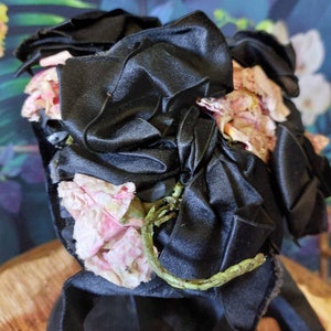 Pu&ograve; includere: Un fiocco di nastro di raso nero con fiori di stoffa rosa. Il fiocco &egrave; legato in un grande nodo e i fiori sono disposti intorno al nodo.