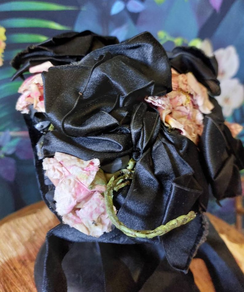 Pu&ograve; includere: Un fiocco di nastro di raso nero con fiori di stoffa rosa. Il fiocco &egrave; legato in un grande nodo e i fiori sono disposti intorno al nodo.