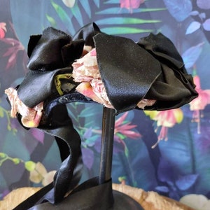 Pu&ograve; includere: Un fiocco di nastro di raso nero con fiori di tessuto rosa. Il fiocco &egrave; su un supporto nero.