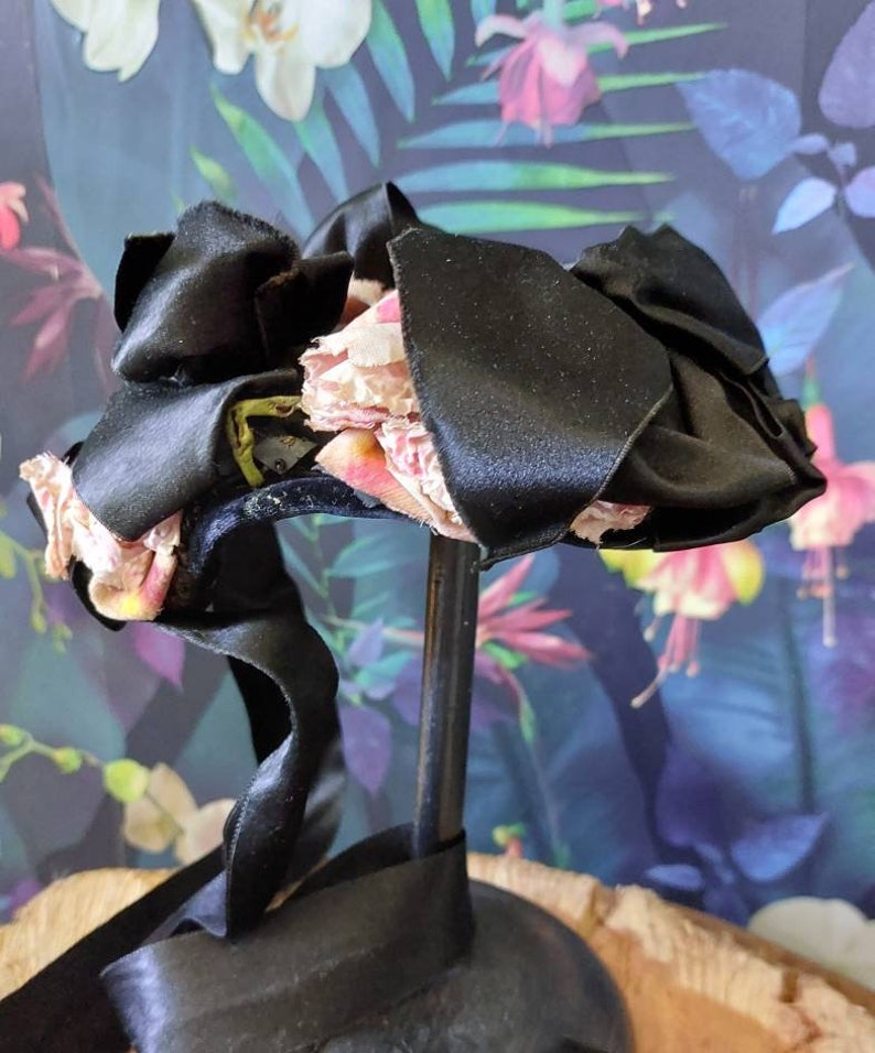 Pu&ograve; includere: Un fiocco di nastro di raso nero con fiori di tessuto rosa. Il fiocco &egrave; su un supporto nero.