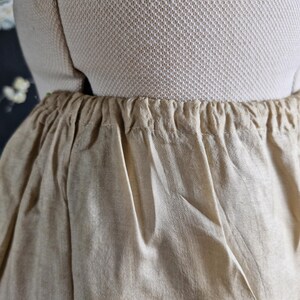 May include: Close-up of beige linen pants with an elastic waistband. The fabric is slightly wrinkled, and the pants appear to be lightweight. The garment is displayed on a mannequin, showcasing the gathered waist detail.