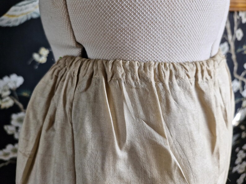 May include: Close-up of beige linen pants with an elastic waistband. The fabric is slightly wrinkled, and the pants appear to be lightweight. The garment is displayed on a mannequin, showcasing the gathered waist detail.
