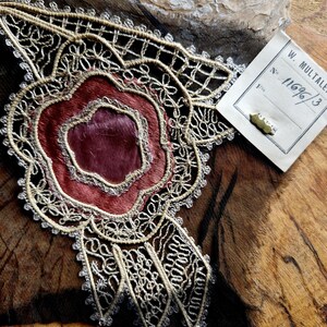 May include: A close-up of a delicate, cream-colored lace trim with a burgundy velvet center. The lace has a floral design and is attached to a piece of brown leather. A white tag with the text "W. Multaier No. 1169/3" is visible in the upper right corner.