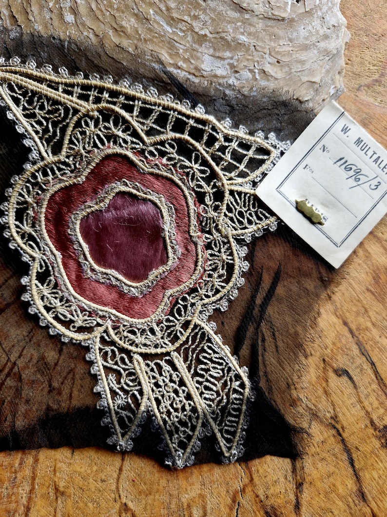 May include: A close-up of a delicate, cream-colored lace trim with a burgundy velvet center. The lace has a floral design and is attached to a piece of brown leather. A white tag with the text "W. Multaier No. 1169/3" is visible in the upper right corner.