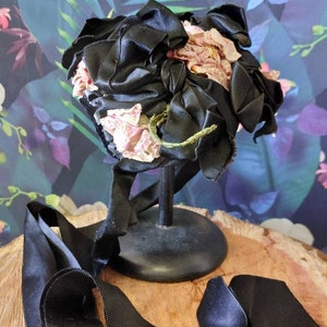 Pu&ograve; includere: Un cappello di raso nero con un grande fiore rosa come ornamento. Il cappello &egrave; su un supporto nero e un nastro di raso nero &egrave; appoggiato su una superficie di legno.