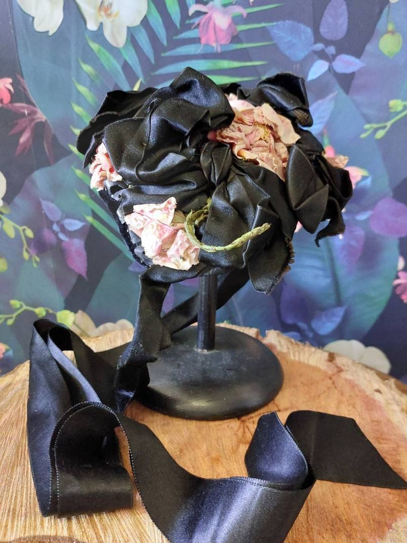 Pu&ograve; includere: Un cappello di raso nero con un grande fiore rosa come ornamento. Il cappello &egrave; su un supporto nero e un nastro di raso nero &egrave; appoggiato su una superficie di legno.