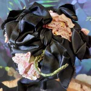 Pu&ograve; includere: Un cappello di raso nero con fiori di stoffa rosa. Il cappello &egrave; su un supporto nero.
