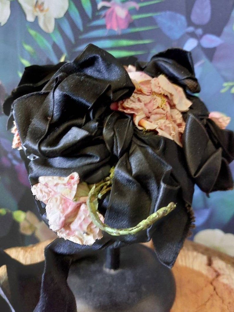Pu&ograve; includere: Un cappello di raso nero con fiori di stoffa rosa. Il cappello &egrave; su un supporto nero.