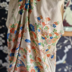 May include: A cream-colored, vintage kimono-style top with a vibrant floral pattern in orange, blue, and green. The design includes stylized flowers, leaves, and a flowing river scene. The top is displayed on a mannequin.