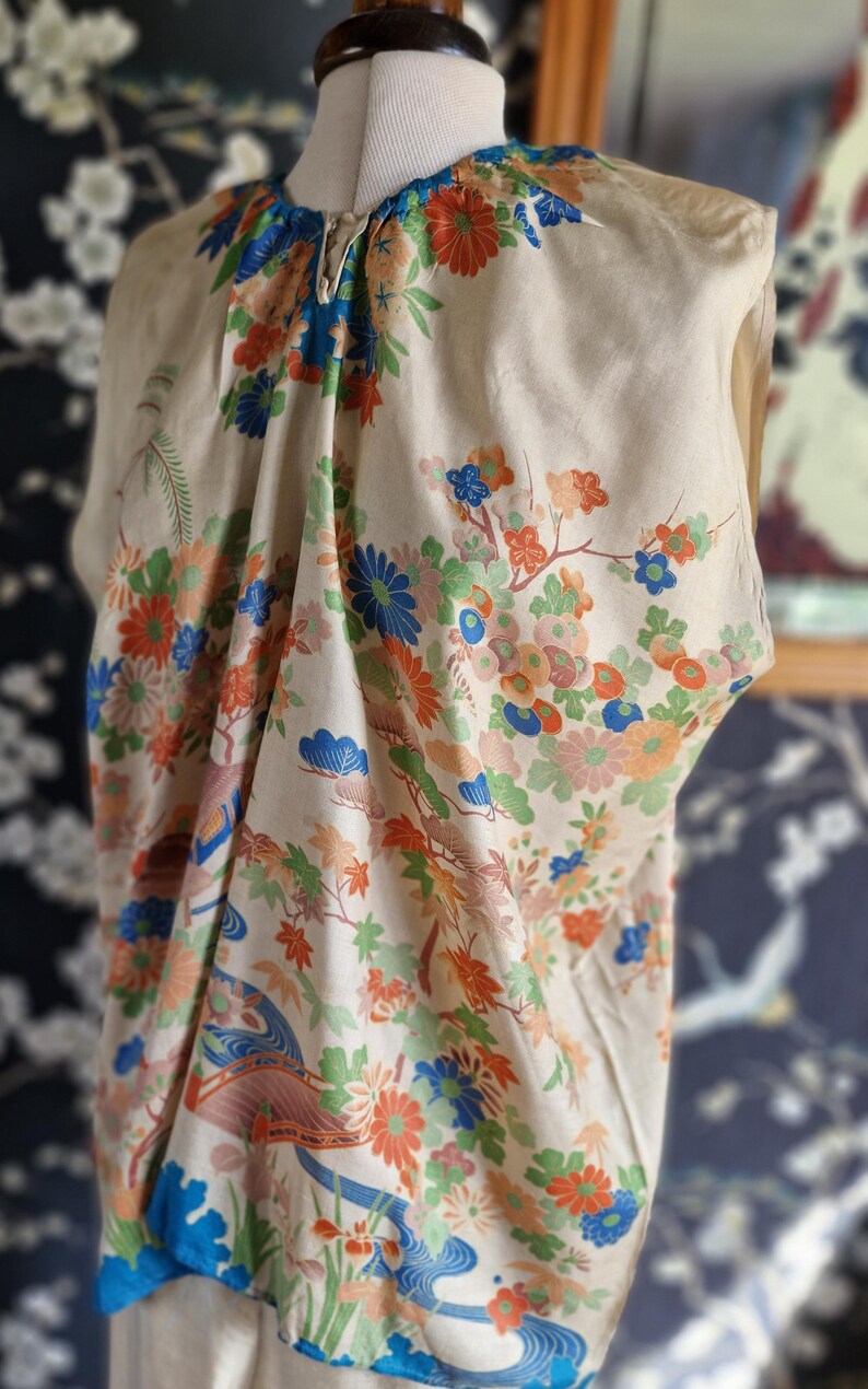 May include: A cream-colored, vintage kimono-style top with a vibrant floral pattern in orange, blue, and green. The design includes stylized flowers, leaves, and a flowing river scene. The top is displayed on a mannequin.
