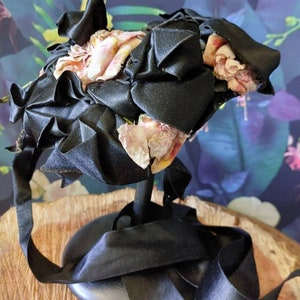 Pu&ograve; includere: Un cappello di raso nero con fiori di stoffa rosa e gialli. Il cappello &egrave; su un supporto nero con un nastro nero.