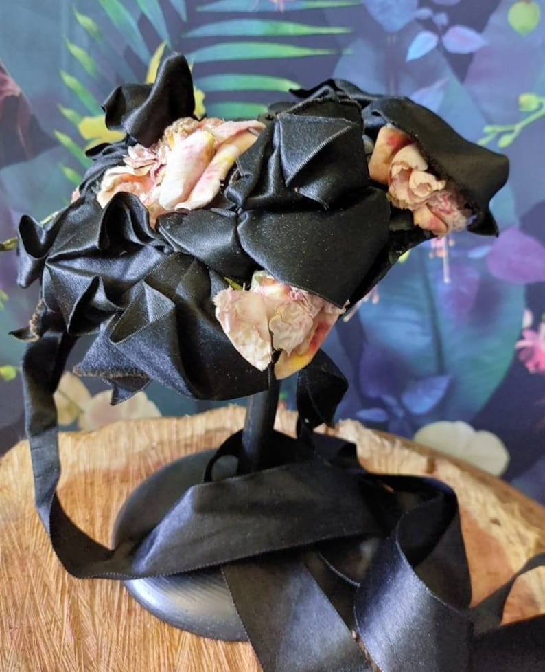 Pu&ograve; includere: Un cappello di raso nero con fiori di stoffa rosa e gialli. Il cappello &egrave; su un supporto nero con un nastro nero.