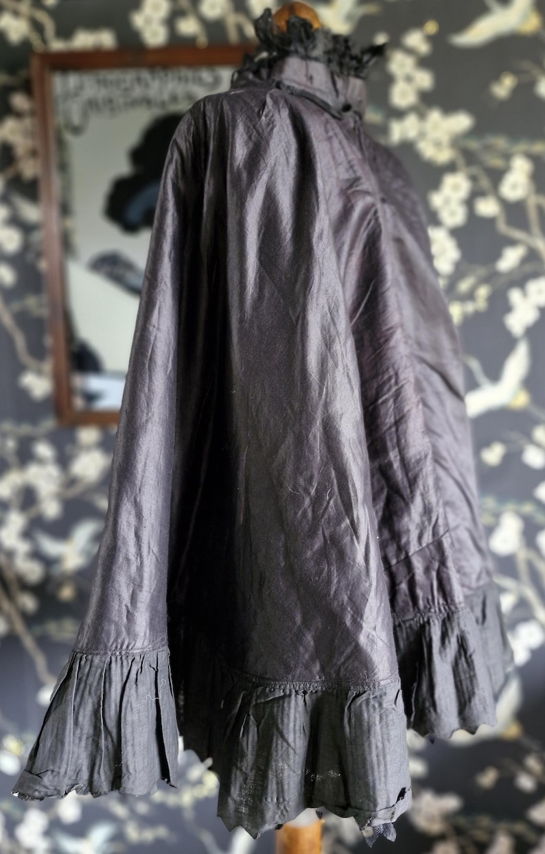 May include: A black silk cape with a ruffled trim. The cape is draped over a mannequin and has a high collar.