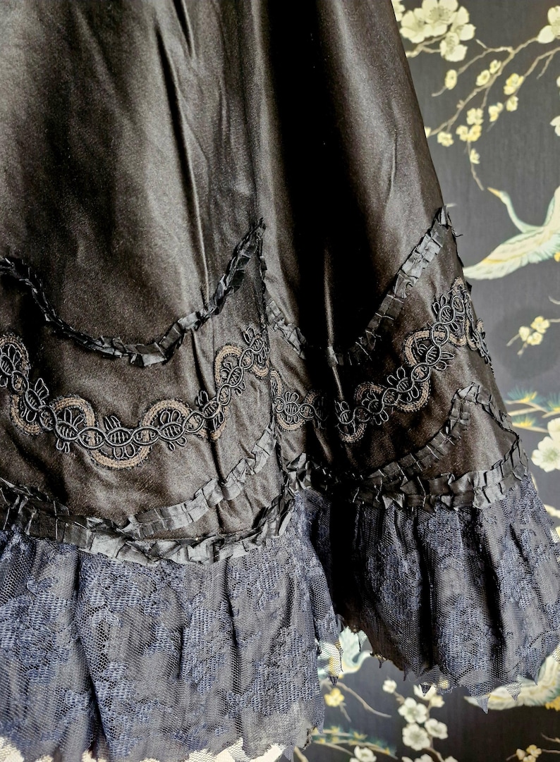 May include: Black satin skirt with a layered ruffle trim and black lace edging. The skirt has a scalloped design with a delicate black embroidery.