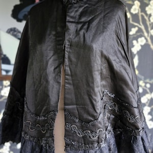 May include: A black satin cape with a lace trim and a high collar. The cape is on a mannequin and has a center front opening.