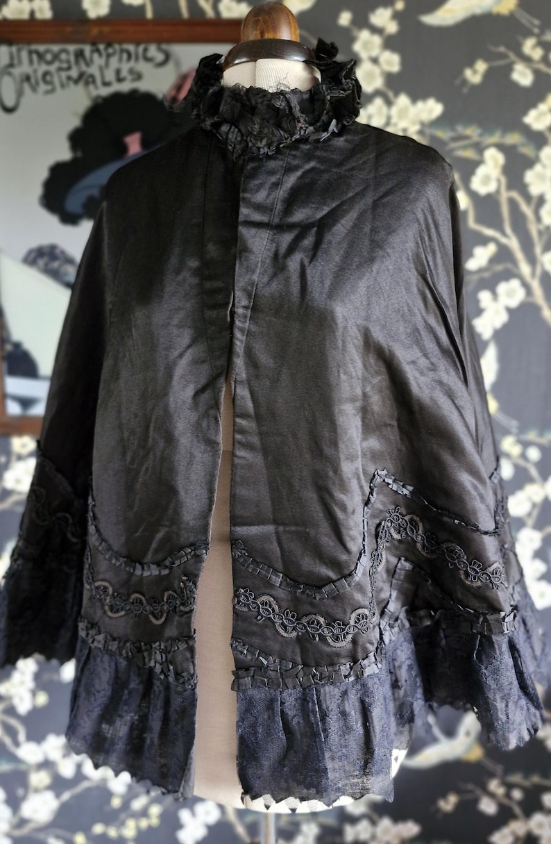 May include: A black satin cape with a lace trim and a high collar. The cape is on a mannequin and has a center front opening.
