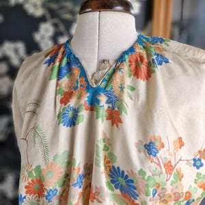 May include: A cream-colored top with a V-neckline, adorned with a vibrant floral pattern in shades of orange, blue, and green. The fabric is draped over a mannequin, showcasing the garment's design and fit. The floral print includes detailed leaves and blossoms.