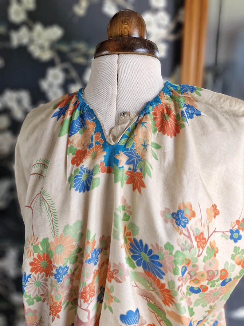 May include: A cream-colored top with a V-neckline, adorned with a vibrant floral pattern in shades of orange, blue, and green. The fabric is draped over a mannequin, showcasing the garment's design and fit. The floral print includes detailed leaves and blossoms.