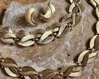 May include: A vintage gold-tone jewelry set featuring a necklace and matching clip-on earrings. The necklace and earrings have a leaf-like design with a textured finish. The jewelry is displayed on a neutral-toned surface.