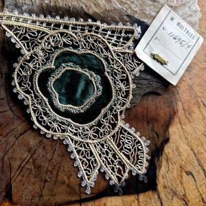 May include: A gold and silver lace trim with a green velvet center. The trim has a floral design and is attached to a piece of black fabric. The trim is labeled with a tag that reads "W. Multaler No. 11696/4".
