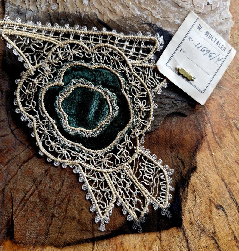May include: A gold and silver lace trim with a green velvet center. The trim has a floral design and is attached to a piece of black fabric. The trim is labeled with a tag that reads "W. Multaler No. 11696/4".