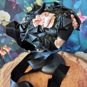 Pu&ograve; includere: Un cappello di nastro di raso nero con fiori di tessuto rosa e paillettes nere.