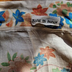 May include: Close-up of a beige fabric with a floral pattern in blue, orange, and green. The fabric has a tag that reads "MADE IN JAPAN". The floral design includes stylized leaves and flowers.