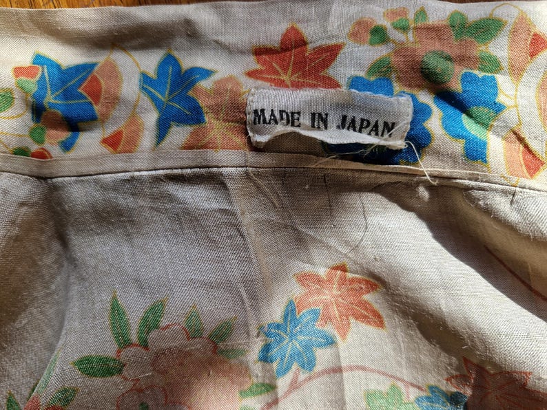 May include: Close-up of a beige fabric with a floral pattern in blue, orange, and green. The fabric has a tag that reads "MADE IN JAPAN". The floral design includes stylized leaves and flowers.