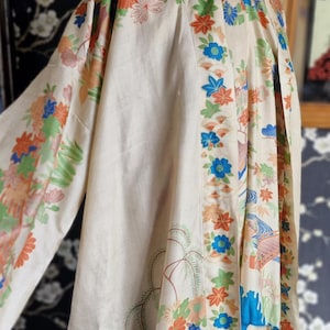 May include: A cream-colored kimono with a floral pattern in orange, blue, and green. The kimono features long sleeves and a decorative border with stylized flowers and landscape elements. The fabric appears to be lightweight.