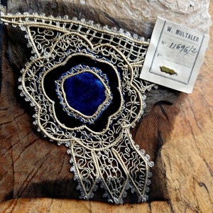 May include: A close-up of a gold and silver lace trim with a blue velvet center. The trim is on a black fabric background. A tag with the text "W. M&Uuml;LTALER No. 11696/2" is visible in the upper right corner of the image.